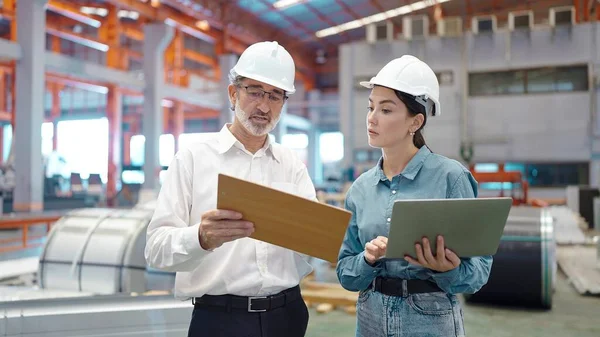 Two engineer manager leader and woman assistant holding laptop wearing helmet talking and checking production standing in workplace area at manufacturing factory