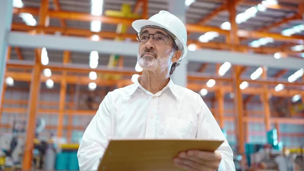 Mühendis başı kask takıyor elinde pano duruyor ve imalat fabrikasına bakıyor iş yerindeki üretim stoklarını tekrar kontrol ediyor.