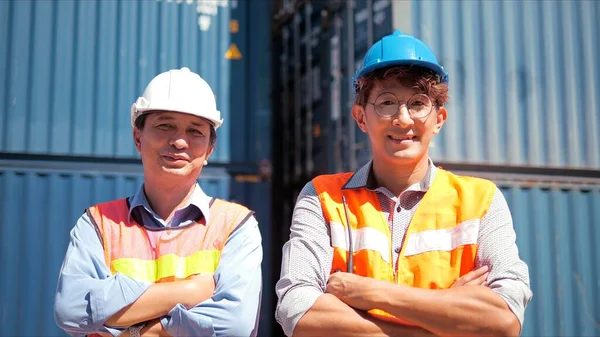 Two professional Asian engineers logistics business in protective helmets crossing arms standing in warehouse cargo container background. They looking confidently