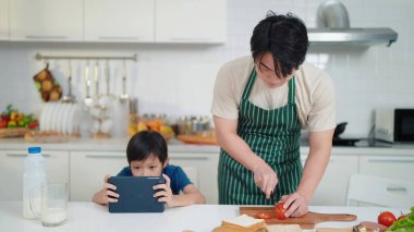 Asyalı baba küçük oğluna kahvaltı hazırlamak için domates kesiyor. Oğlum mutfakta tatilde oturup tabletle oyun oynuyor.