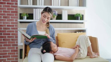 Mutlu Asyalı anne, evde aile hayatı hobisinin keyfini çıkarırken kanepeye uzanan kızına masal kitabı okuyor. Mutlu aile kavramı, birlikte zaman geçirmek.