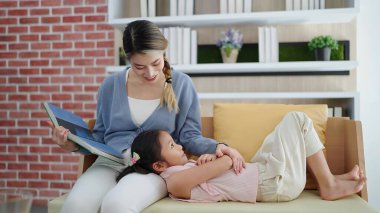 Mutlu Asyalı anne, evde aile hayatı hobisinin keyfini çıkarırken kanepeye uzanan kızına masal kitabı okuyor. Mutlu aile kavramı, birlikte zaman geçirmek.