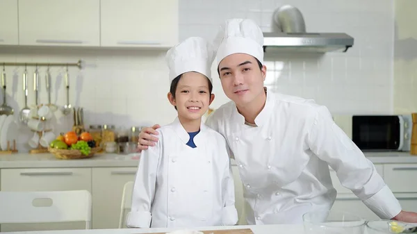 シェフの衣装を着た幸せなアジアの父と小さな息子が笑顔で 自宅のキッチンルームでカメラを見ています 幸せな家庭のコンセプト — ストック写真