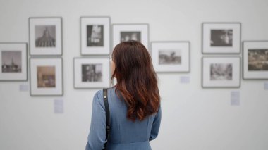 Düşünceli kadın galeri ziyaretçisi sergi salonunda duvarın yanında duruyor, resim ve sanat eserlerine bakıyor.