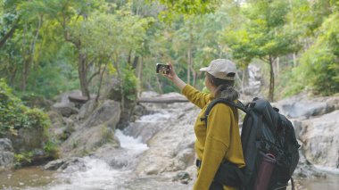 Asyalı genç bir kadın yürüyüş yapıyor, sırt çantası giyiyor, sarı kazak giyiyor ve ormanda akıllı telefon görüşmeleri yapıyor.