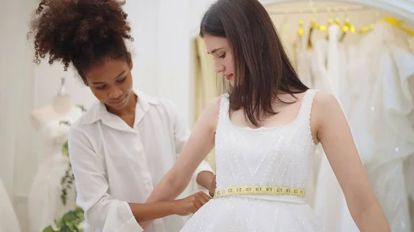 若いアフリカ系アメリカ人女性ファッションデザイナー使用してテープメジャーサイズの美しいアジアの花嫁で白いウェディングドレスでウェディングスタジオ — ストック写真
