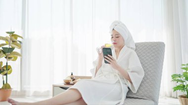 Bornozlu ve havlu sarıklı güzel bir Asyalı kadın şampanya içerken modern oturma odasındaki kanepede dinlenirken akıllı telefon kullanıyor.