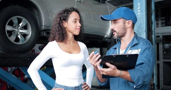 車の修理店で女性のクライアントへの車のメンテナンスや修理を説明する統一されたチェックリストの自動車整備士 車のサービス メンテナンス ジェスチャーや男性の機械的コンセプト — ストック写真