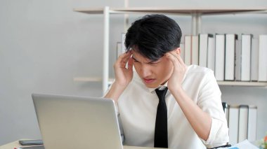 Mutsuz Asyalı genç adam çalışırken endişeli bir yüz ve alnı olan dizüstü bilgisayara bakıyor. Migren ağrısı çeken bir iş adamı.