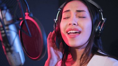 Genç Asyalı kadın sanatçı müzik stüdyosunda şarkı söylüyor. Mikrofon kullanarak kulaklık takan kadın şarkıcı neon stüdyoda şarkı söylüyor.