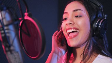 Genç Asyalı kadın sanatçı müzik stüdyosunda şarkı söylüyor. Mikrofon kullanarak kulaklık takan kadın şarkıcı neon stüdyoda şarkı söylüyor.
