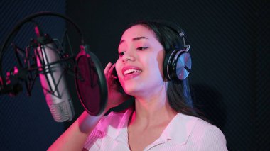 Genç Asyalı kadın sanatçı müzik stüdyosunda şarkı söylüyor. Mikrofon kullanarak kulaklık takan kadın şarkıcı neon stüdyoda şarkı söylüyor.