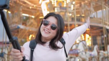 Asyalı bayan gezgin, eğlence parkında tatilin yapılacağı yeri göstermek için neşeli hareketli fotoğraf makinesiyle selfie çekiyor.