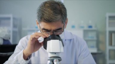 Yüz maskesi takan Asyalı doktor, aşı örneğini laboratuvar ve aşı araştırmalarında mikroskop ile analiz ediyor. Karantina veya virüs salgını kavramı. 