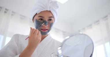 Yüzüne gri maske takan Asyalı bir kadın. Saçları beyaz havluya sarılı. Kızlar için güzellik merkezi prosedürleri. Cilt bakımı, nemlendirici ve nemlendirici kavram.