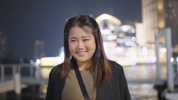 アジアの女性は笑顔でカメラを見ながら 夜には都会の近代的な街で一人で屋外に立って幸せ — ストック動画