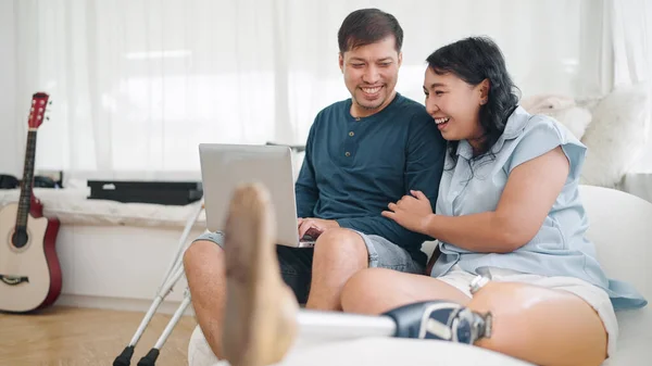 Happy asian woman in leg prosthesis and her husband sitting on comfortable sofa while typing on laptop for working online or chatting on social media at home. Leg prosthetic equipment