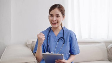 Gülümseyen genç Asyalı doktor ya da üniformalı hemşire steteskoplu elinde tabletle kameraya bakıp elini kaldırıyor. Hasta ile görüntülü konuşma yaparken Asyalı kadın doktor kameraya bakıyor.