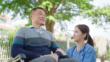 Asian woman caregiver nurse with elderly male patient in wheelchair at outdoors. Caregiver nurse take care of elderly mature man patient in wheelchair at public park