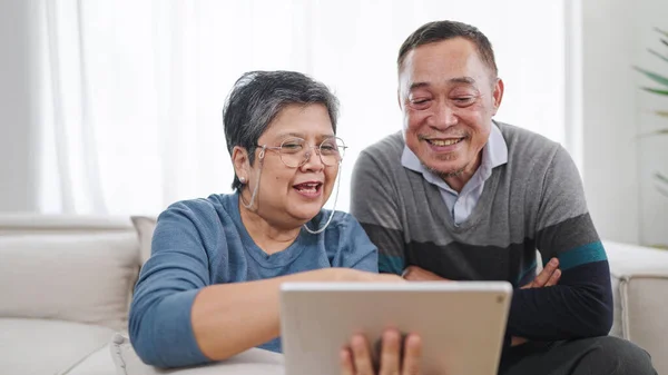 幸せな年配のアジアのカップルは一緒に話し タブレットを使用して自宅のリビングルームに座っている間 ソーシャルメディアを閲覧を楽しんでいます 高齢のカップルは 瞬間を愛しています カップル定年生活 — ストック写真