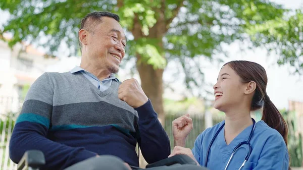Asian woman caregiver nurse cheer up to elderly male patient sitting in wheelchair. Caregiver nurse take care and support elderly male patient