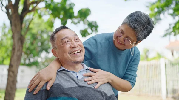 Happy asian elderly married couple relaxing in park together. Cheerful asian adult male patient sitting on wheelchair and senior wife enjoying leisure. Relationship concept