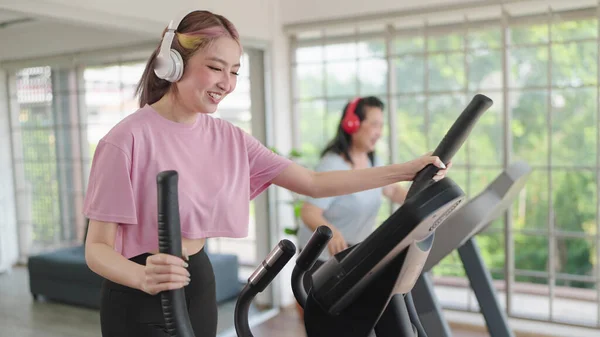 Happy young asian women in sportswear wearing headphones while exercising on the machine at home. Young women talking with mother while training on elliptical machine. Health care concept