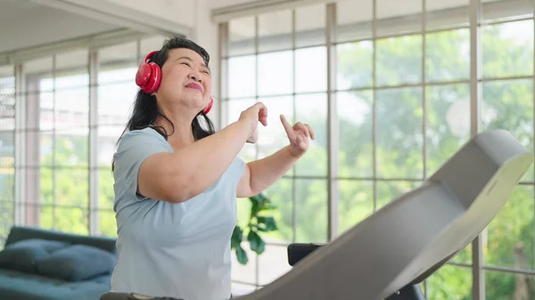 Happy asian elderly women in headphones enjoy dancing while training on elliptical machine at home. Elderly women exercising on the machine. Elderly activity, Health care concept