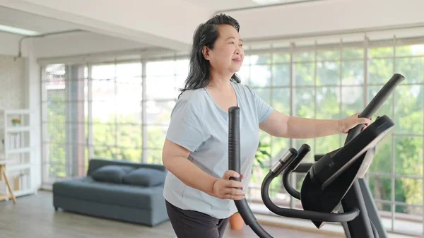 Asian elderly women exercising on the machine in living room at home. Mature woman enjoy doing exercising on treadmill for well being slimming weight. Elderly lifestyle concept
