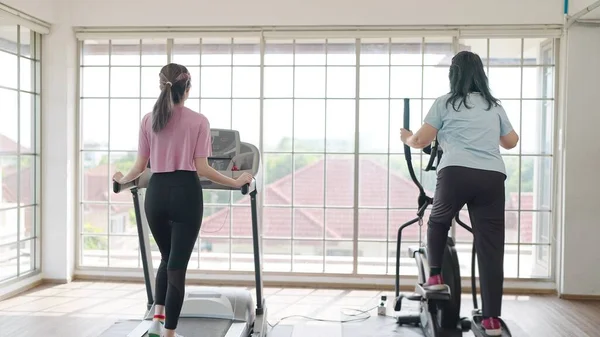 Back view of asian elderly mother and daughter enjoy exercising on the machine in living room at home on weekend. Elderly mother and daughter talking together while exercising on the machine