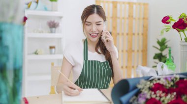 Önlüklü güzel Asyalı kadın çiçekçi. Akıllı telefondan konuşuyor. Müşteriyle sipariş veriyor ve deftere yazı yazıyor. Küçük işletme kavramı