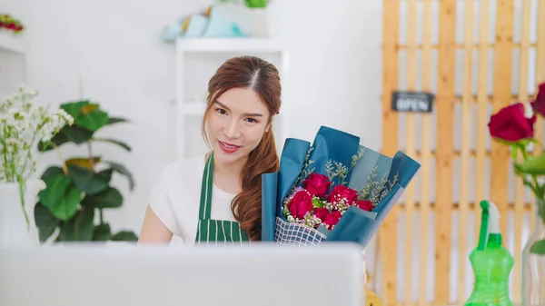 Young asian women owner floral store holding bouquet of rose flowers on hands while video call on laptop communicating with client. Selling bouquet through online live streaming on laptop