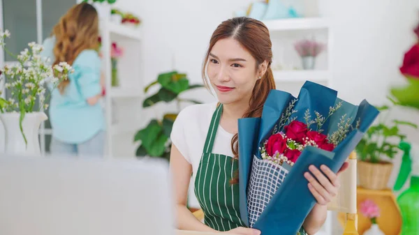 Young asian women owner floral store holding bouquet of rose flowers on hands while video call on laptop communicating with client. Selling bouquet through online live streaming on laptop
