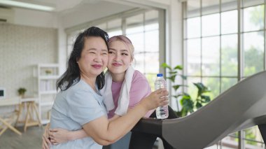 Evdeki makinede egzersiz yapan güzel Asyalı kadınlar. Yaşlı kadınlar, eliptik makinede çalışırken kızlarına su veriyorlar. Mutlu aile, sağlık hizmeti kavramı