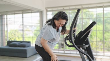 Evdeki oturma odasındaki makinede egzersiz yapmaktan bacakları ya da dizleri ağrıyan Asyalı yaşlı kadınlar. Yaşlılık, sağlık sorunları ve insanların konsepti. Egzersiz yüzünden ağrı.