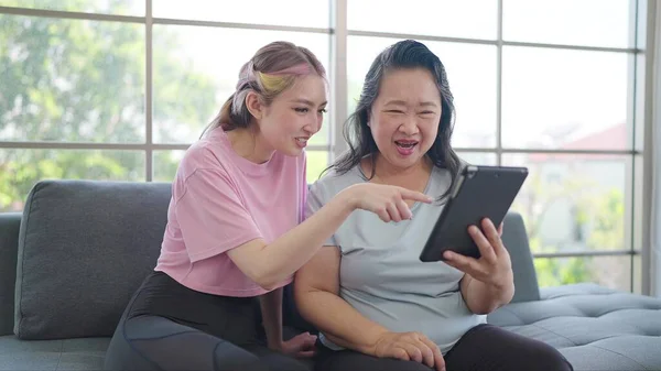 Young daughter and elderly mother enjoy using tablet while sitting on couch in living room at home. Elderly Asian mother and daughter enjoy shopping online on tablet. Happy Asian family