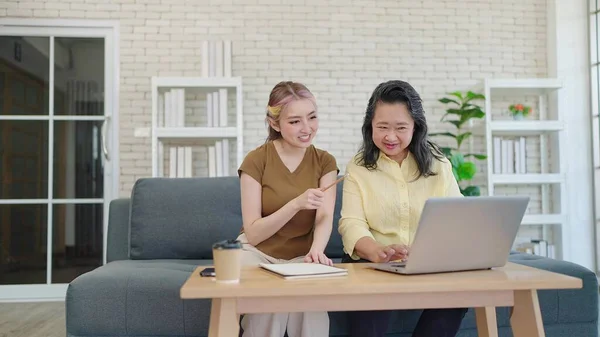 Young asian daughter teaching elderly mother using laptop sitting on couch in living room at home. Mother and daughter using laptop together. Learning how to use modern technologies