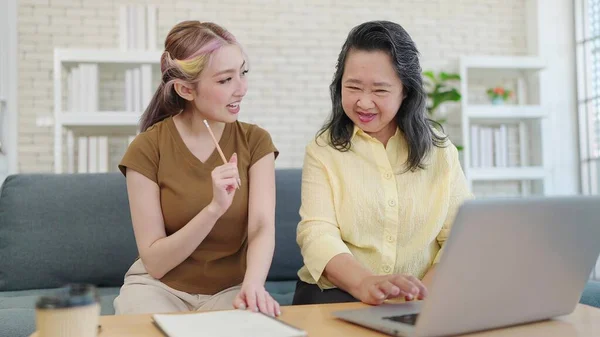 Elderly Asian mother and young daughter using laptop computer together in living room at home. Mother and daughter shopping through e-commerce website on laptop and taking note in notebook