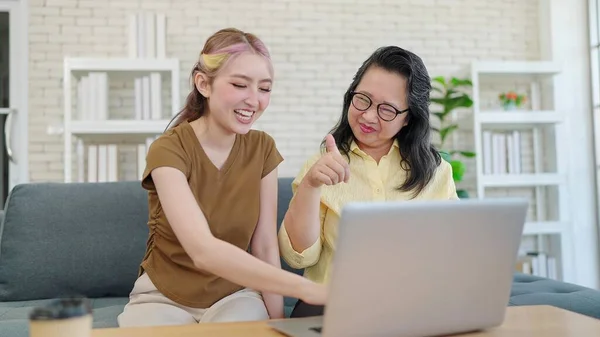 Elderly Asian mother and young daughter using laptop computer together in living room at home. Mother and daughter shopping through e-commerce website on laptop and taking note in notebook