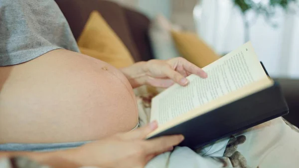 Pregnant woman reading a book while sitting on comfortable sofa in living room at home. Relaxing pregnant woman enjoy reading a book. Pregnancy concept