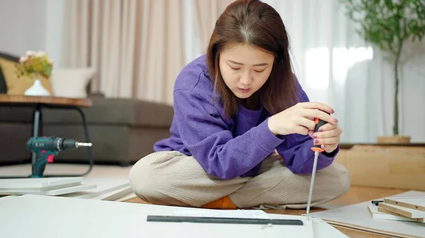 Young Asian woman reading furniture assembly guide on tablet for assembling furniture by herself at home. Woman tightens screw on furniture looking assembly guide on tablet