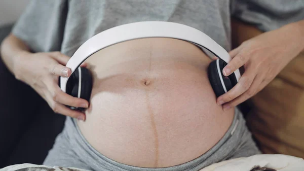 Relaxing pregnant woman holding headphones putting on belly audio therapy unborn baby. Women pregnant putting headphone play music for baby in belly. Pregnancy concept