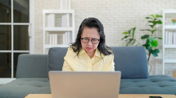 Asian elderly woman in glasses using laptop while sitting on sofa in living room at home. Senior women working or chatting in social media. Retirement lifestyle concept