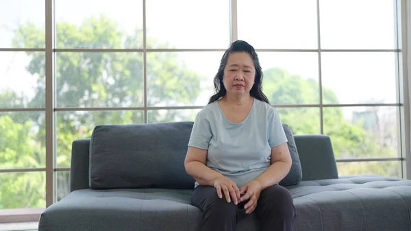 Lonely Asian elderly woman sitting on sofa at home. Unwell senior woman having depression or dizziness while sitting alone in living room at home. Health problem concept