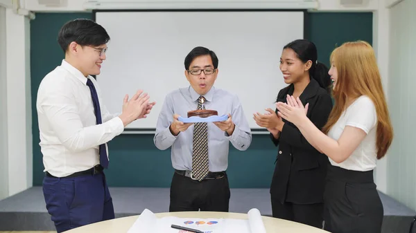Friendly colleagues making present surprise giving birthday cake to Asian man manager while meeting at office. Group of Asian business people celebrating birthday with cake to man colleague
