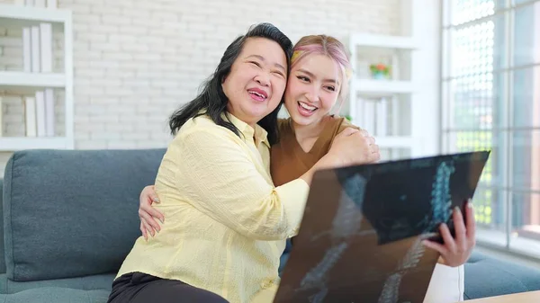 Happy young Asian daughter and mother holding x-ray sheet of bone examination results while sitting on sofa at home. Happy family concept, Healthy concept