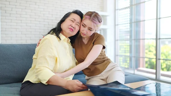 Happy Asian parent mother with young daughter hugging cuddle together while sitting on sofa at home. Young daughter hugging and taking care eldery mother. Happy family concept