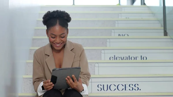 Young Africa America businesswoman using a digital tablet while sitting on the stairs at office. Businesswoman enjoy browsing on tablet while taking a break