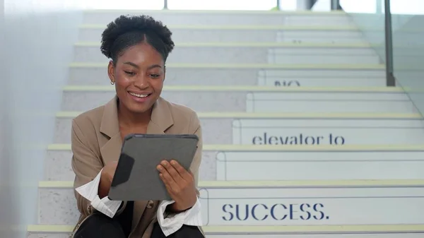 Young Africa America businesswoman using a digital tablet while sitting on the stairs at office. Businesswoman enjoy browsing on tablet while taking a break