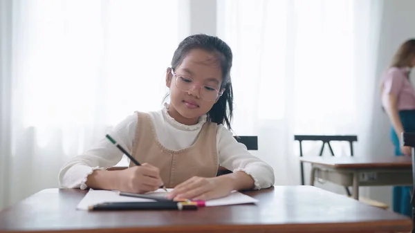 メガネをかけたアジアの女子生徒が教室でノートを書いている 女子高生は試験の準備と勉強をしています 小さな生徒が教室の机で書いている 教育知識コンセプト — ストック写真